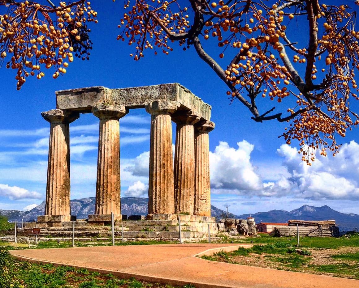 Ancient Greek temple ruins with weathered Doric columns standing against a brilliant blue sky with white clouds. Orange and yellow fruit tree branches frame the top of the image, while mountains are visible in the distance across the Mediterranean landscape.
